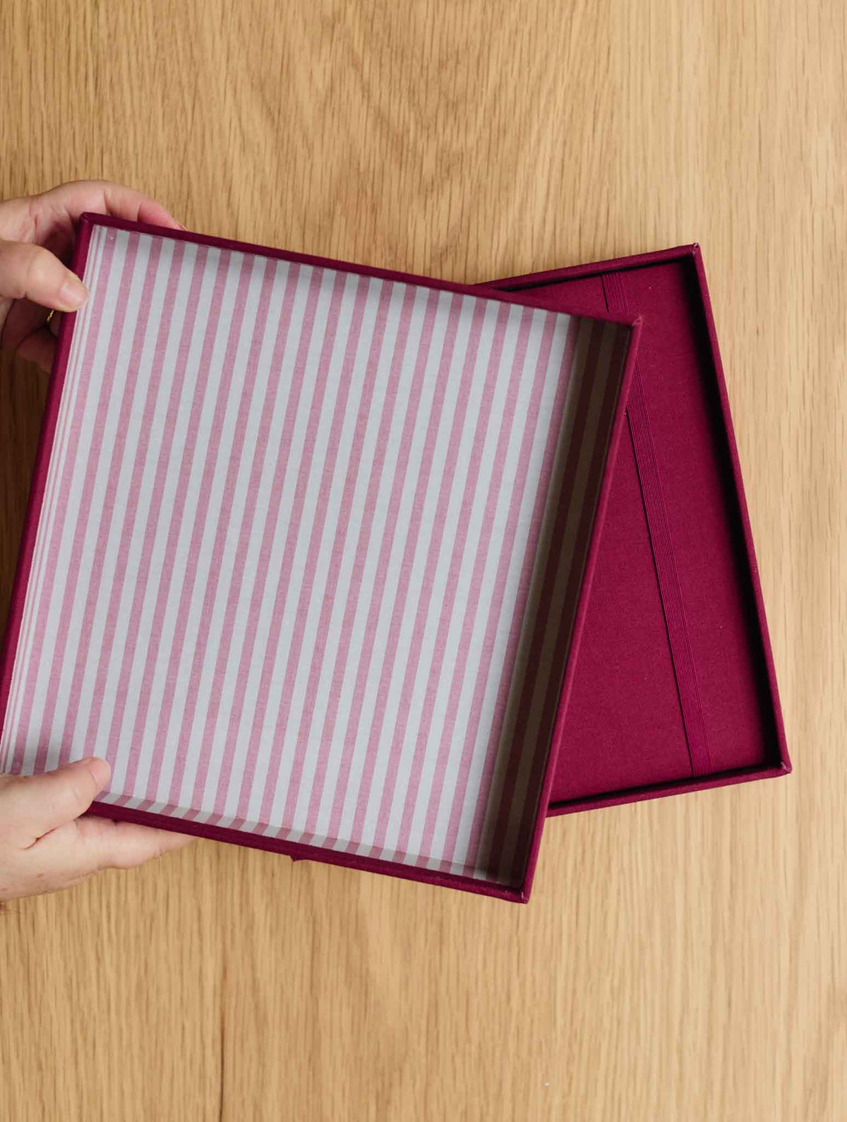 A top-down view of a person's hands holding an open square gift box with a plum exterior and a pink-and-white striped interior, set against a light wood grain background.