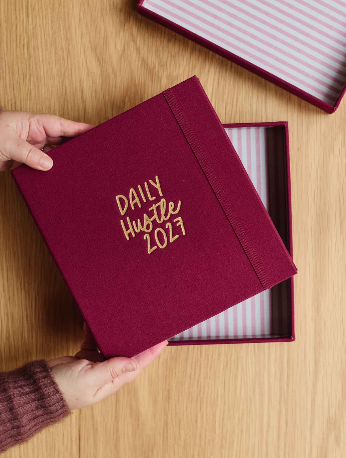 A person's hands are holding a plum linen box with gold foil text reading 'DAILY Hustle 2027' on a light wood background. The box is partially open, revealing a pink and white striped interior.