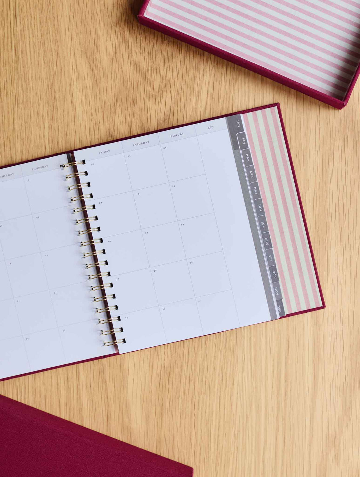 An open spiral-bound planner  on a light wood tabletop, showing a monthly calendar page and daily tabs. A matching pink-and-white striped box is partially visible at the top right.