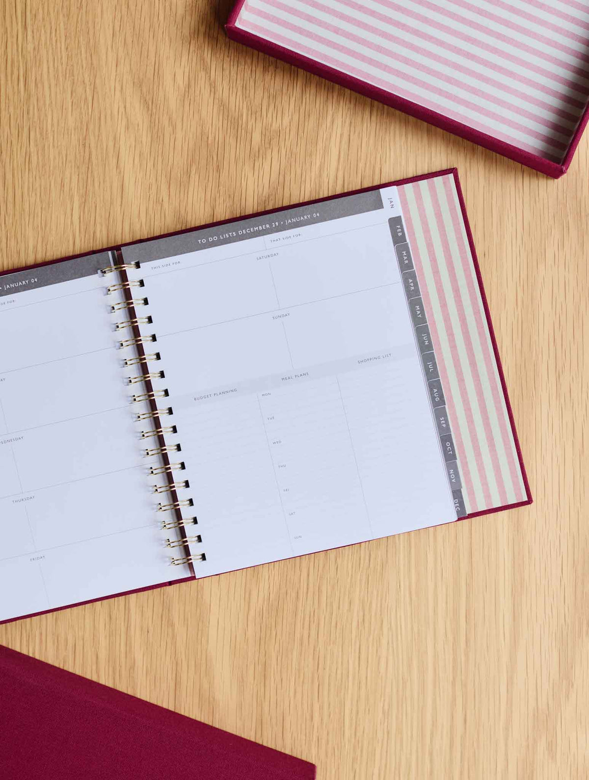An open spiral-bound planner with a plum cover, showing a to do list page laid on a light wood tabletop. A matching plum and pink/white striped box is partially visible at the top right.