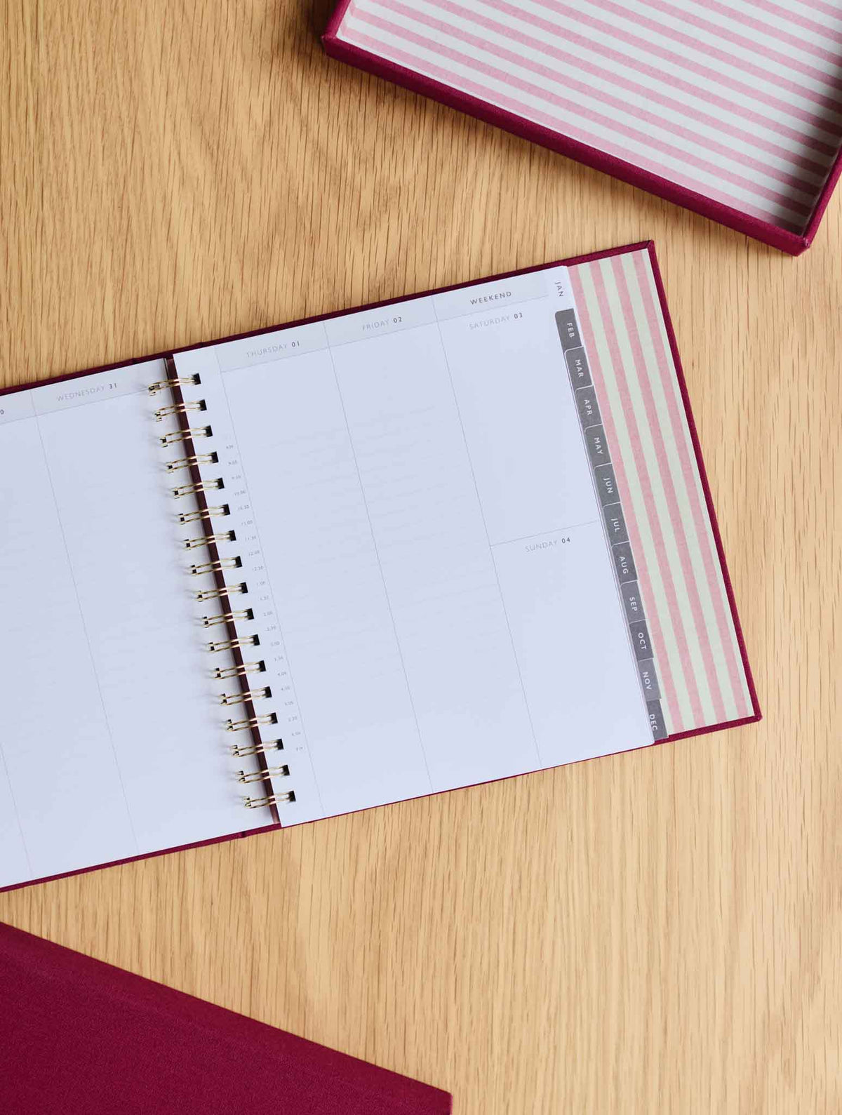 An open weekly planner with white pages and a plum cover lies on a light wood tabletop. A matching box with pink / white striped internal is partially visible at the top right, and the bottom left corner shows the box lid of the same planner in plum.