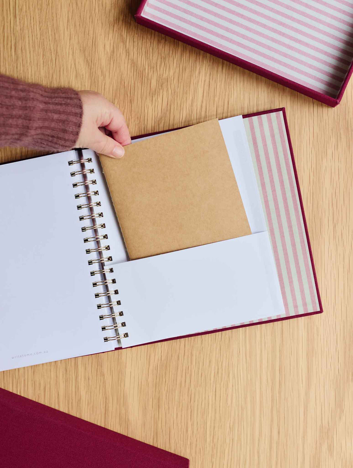 A hand wearing a mauve knit sleeve inserts a brown kraft notebook into pocket of a spiral-bound planner with plum cover, set on a light wood tabletop.