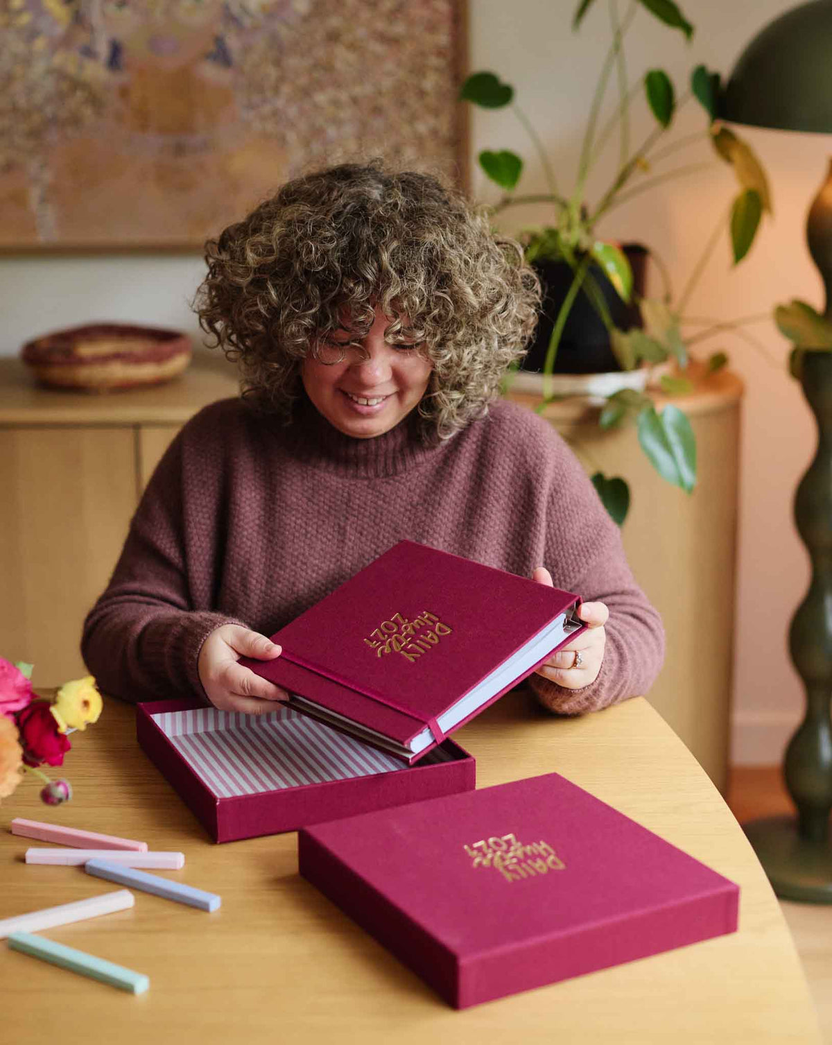 A woman with curly hair sits at a wooden table, smiling as she holds an open plum planner with gold foil lettering. A matching plum box with gold lettering sits open on the table next to her, along with pastel highlighters.
